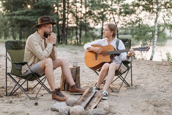 Image Alt Text: A man playing a harmonica and a woman playing a guitar by a campfire