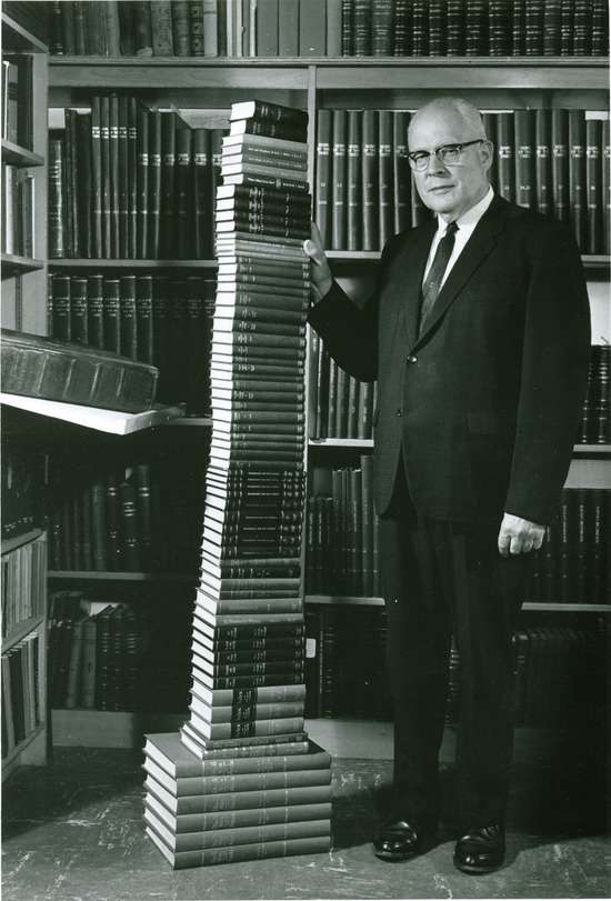 A man standing next to the complete series of Ellen White's writings where each book is stacked on top of each other until the stack is as tall as his head, demonstrating how many books she wrote in her lifetime.