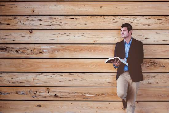 A man leans against a wooden wall while holding his Bible.