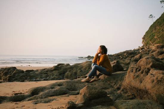A barefoot young woman sits on the rocks at the beach and contemplatively gazes into the sunset