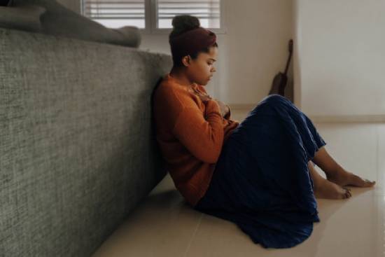 A young woman of color who looks distraught sits on the floor leaning against the back of a couch with a blanket over her legs