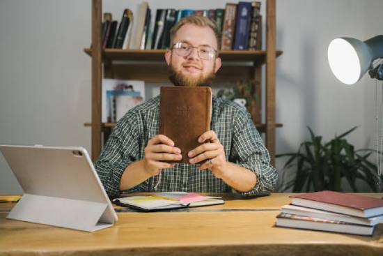 A young man happily sits at a desk holding up his Bible.