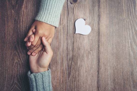 Two hands interlock on a table with a paper cutout heart nearby, indicating love and care between them.