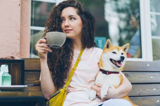 A woman drinks coffee on a neighborhood bench, her dog in her lap.