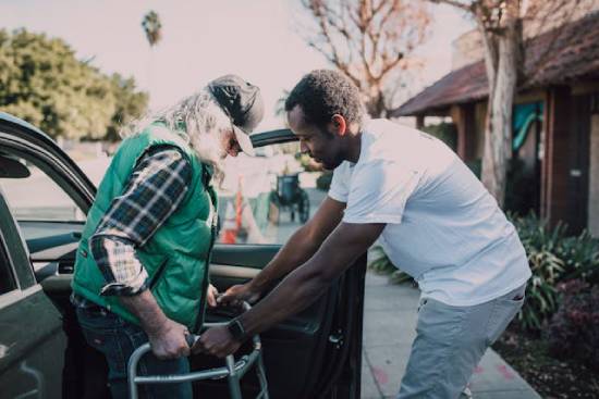 A young man helps an older gentleman with a walker out of his car.