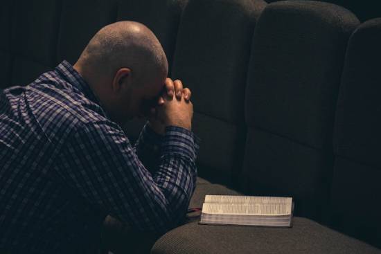Man bowing head and clasping hands in prayer before an open Bible