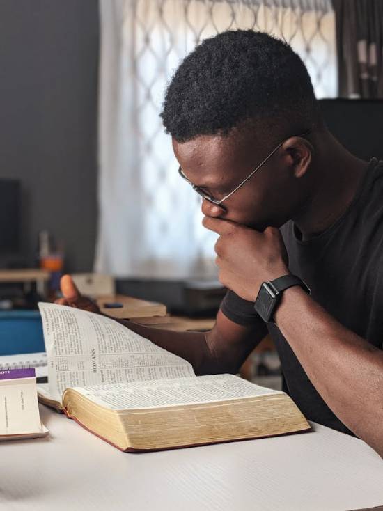a Black man wearing glasses and sitting at a table, with his hand on his chin studying an open Bible, in deep thought