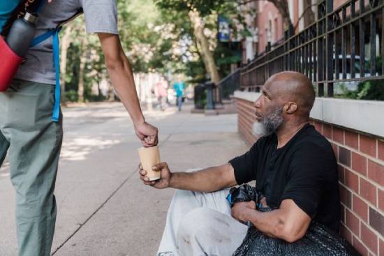 Passerby giving money to a homeless man An unhoused man sits on the sidewalk with a cup as a stranger puts money in it.