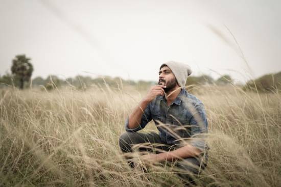 A young man of color squats in tall grass and contemplates something.