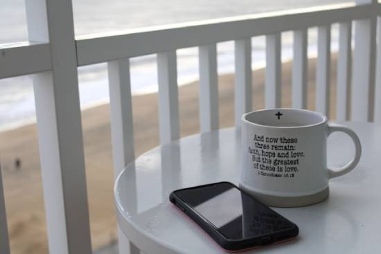 A cellphone and mug with 1 Corinthians 13:13 written out on it sit on a beachfront table.