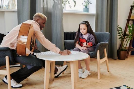 A young man with a guitar in his lap sits at a table and points to notes as he teaches a little girl how to play guitar